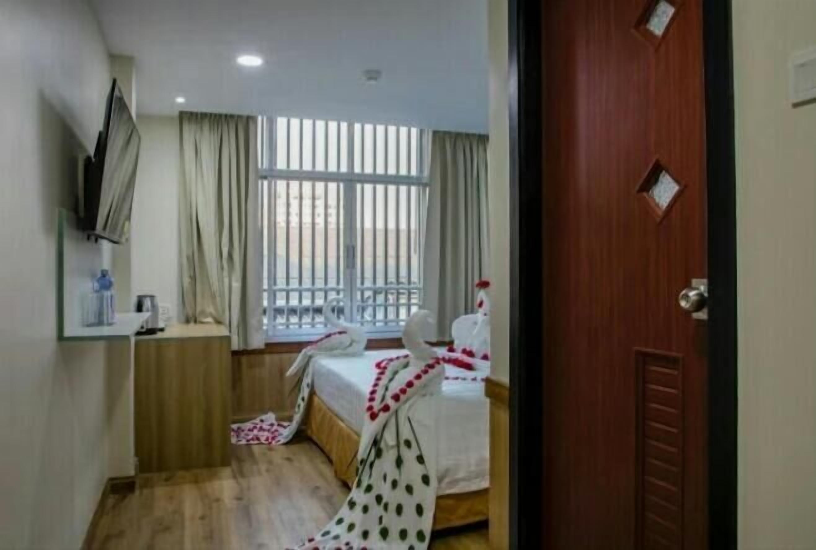 A hotel room with a bed, a TV mounted on the wall, a window with curtains, and a wooden floor.