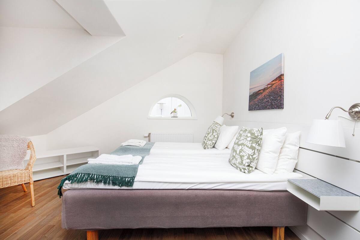 A modern bedroom with a sloped ceiling, a bed with white linens, a green throw blanket, and a view of a cityscape through a window.