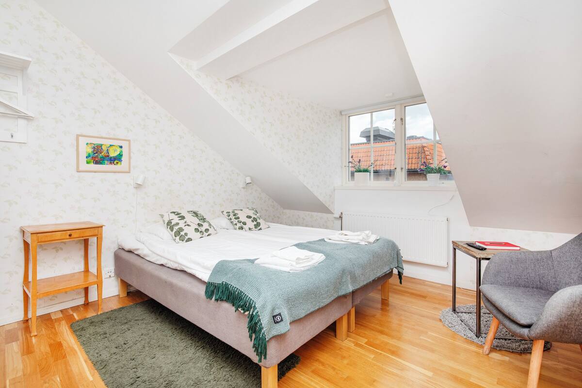 A bedroom with a bed, a small table, a chair, a window, and a radiator.