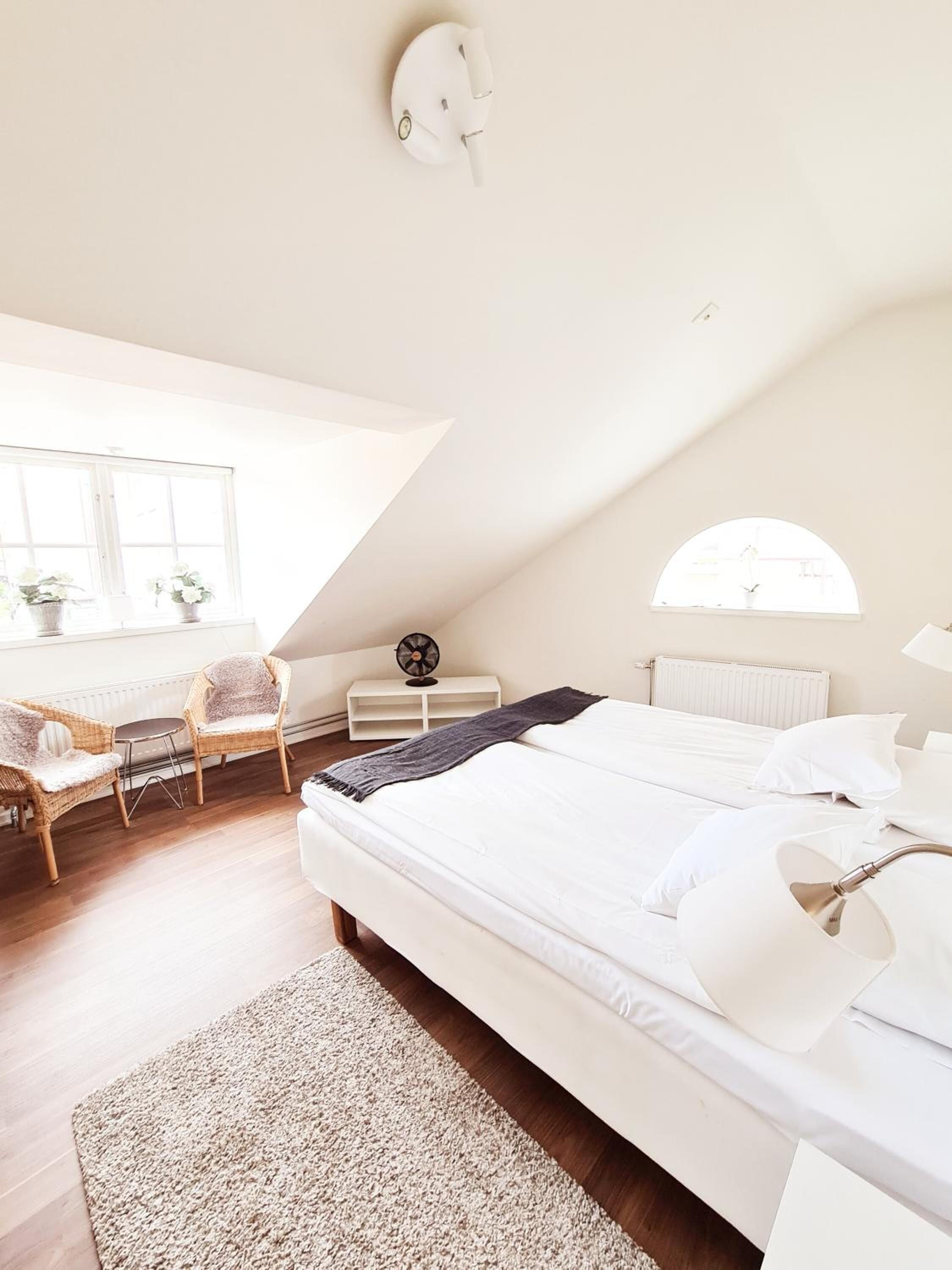 Quarto com uma cama grande, uma escrivaninha, duas cadeiras, um radiador, um ventilador e uma janela.