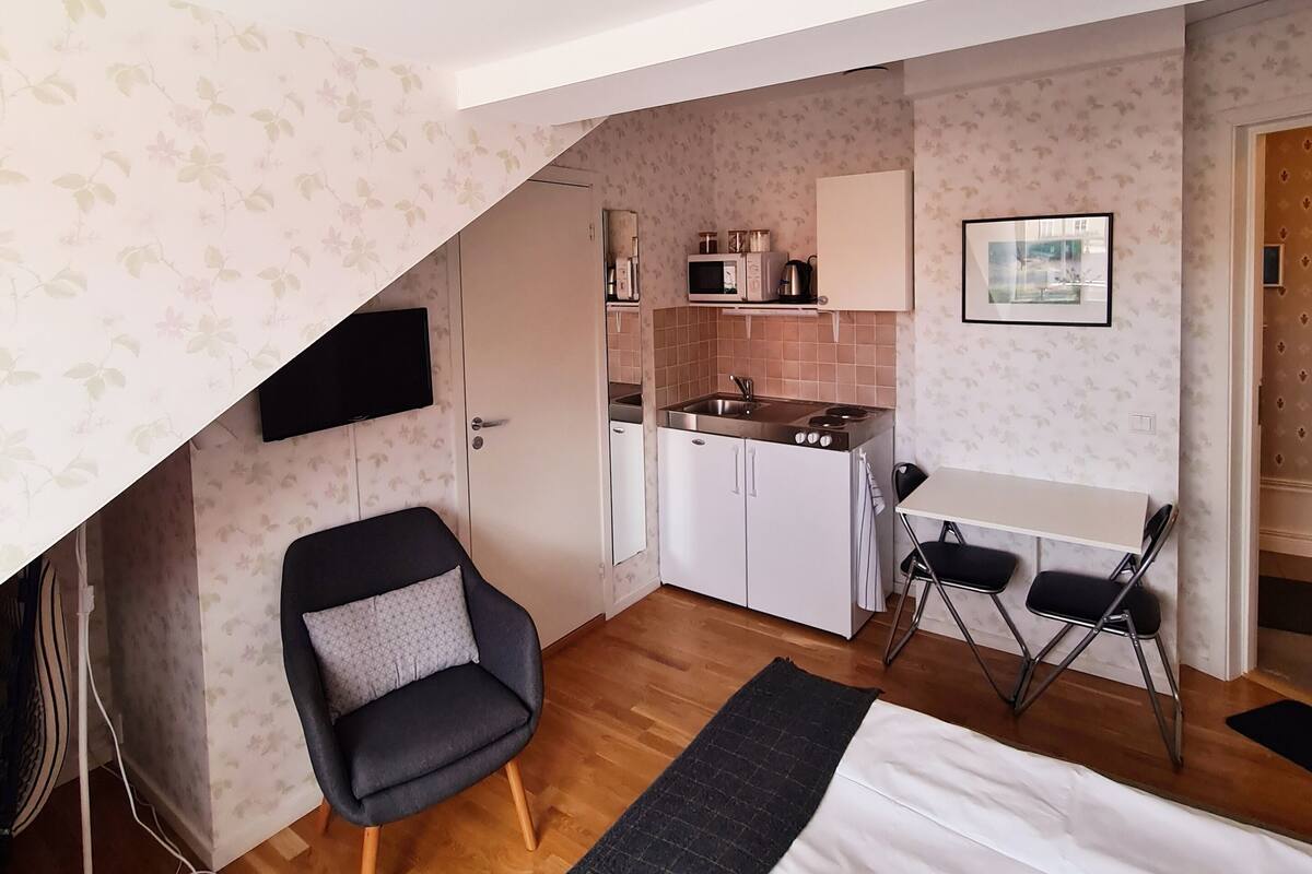 A compact room with a bed, a chair, a small kitchenette, and a TV.