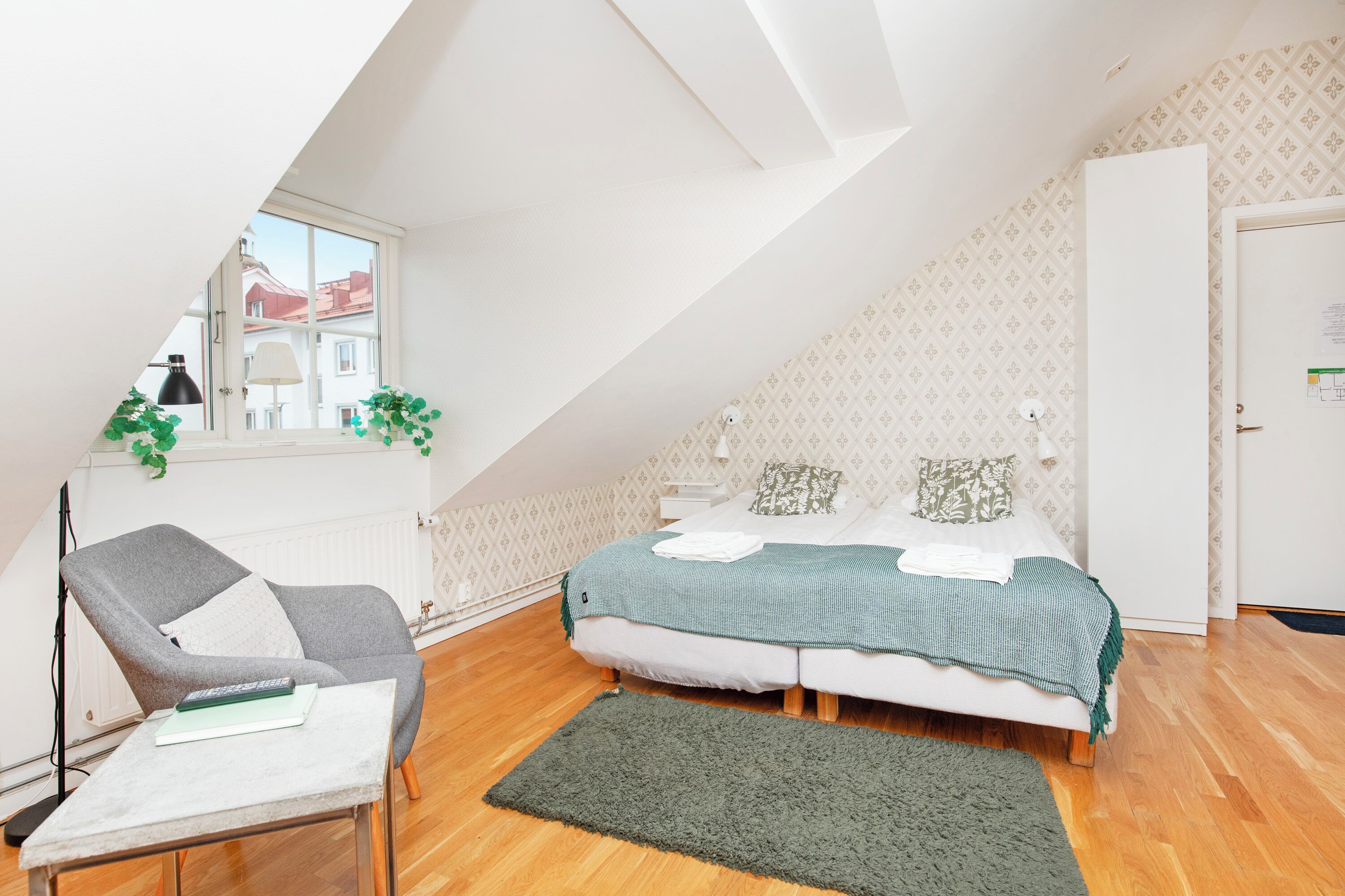Un dormitorio con techo inclinado, una cama con manta verde, un sillón gris, una mesita con un libro y una ventana con vistas a edificios.