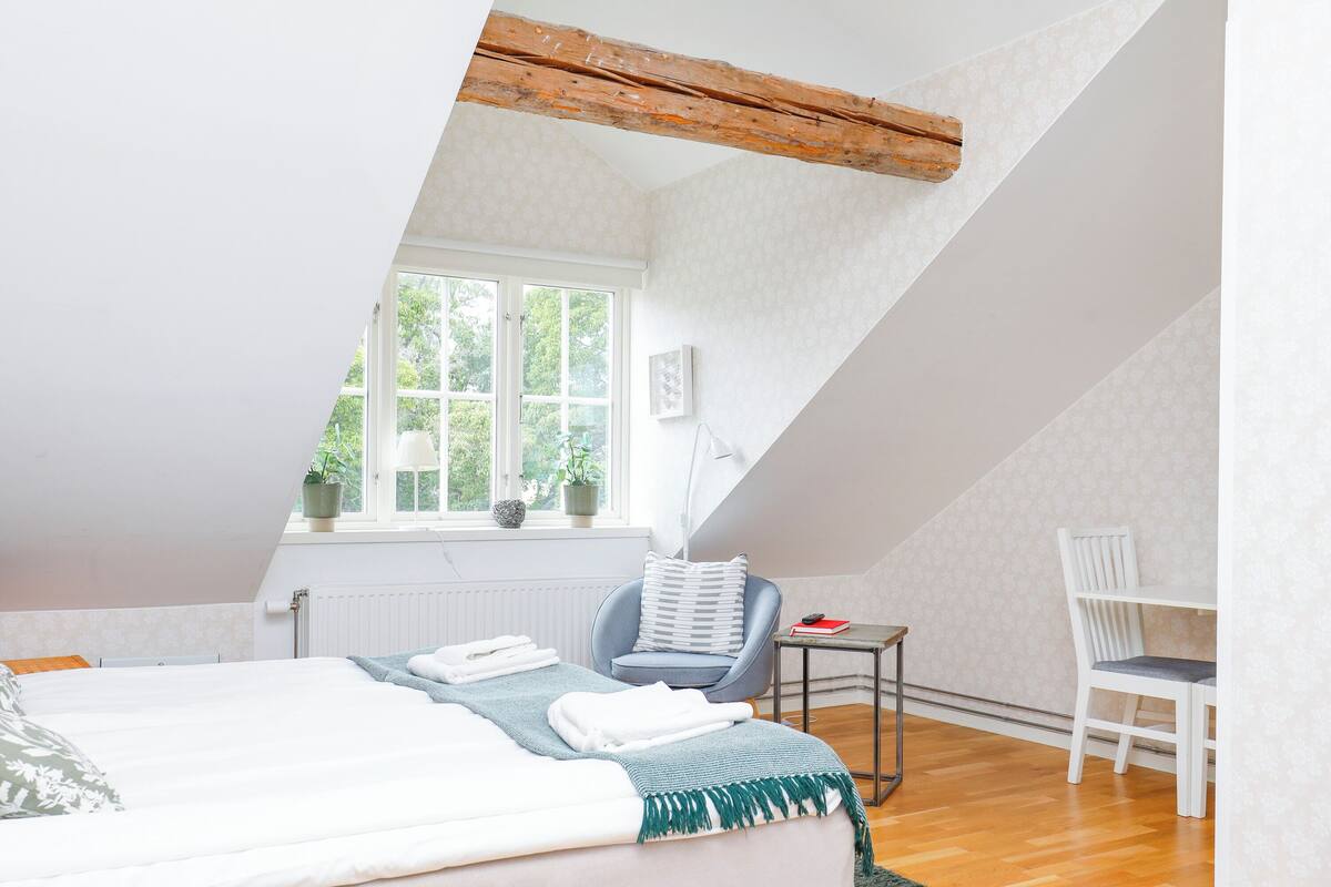 A bedroom with a sloped ceiling, a bed with a green throw, a small table with a book, a chair, and a window with plants.