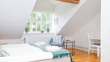 A bedroom with a sloped ceiling, a bed with a green throw, a small table with a book, a chair, and a window with plants.