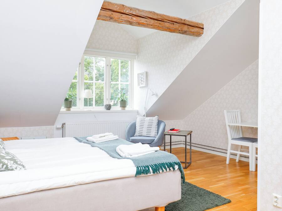 A bedroom with a sloped ceiling, a bed with a green throw, a small table with a book, a chair, and a window with plants.