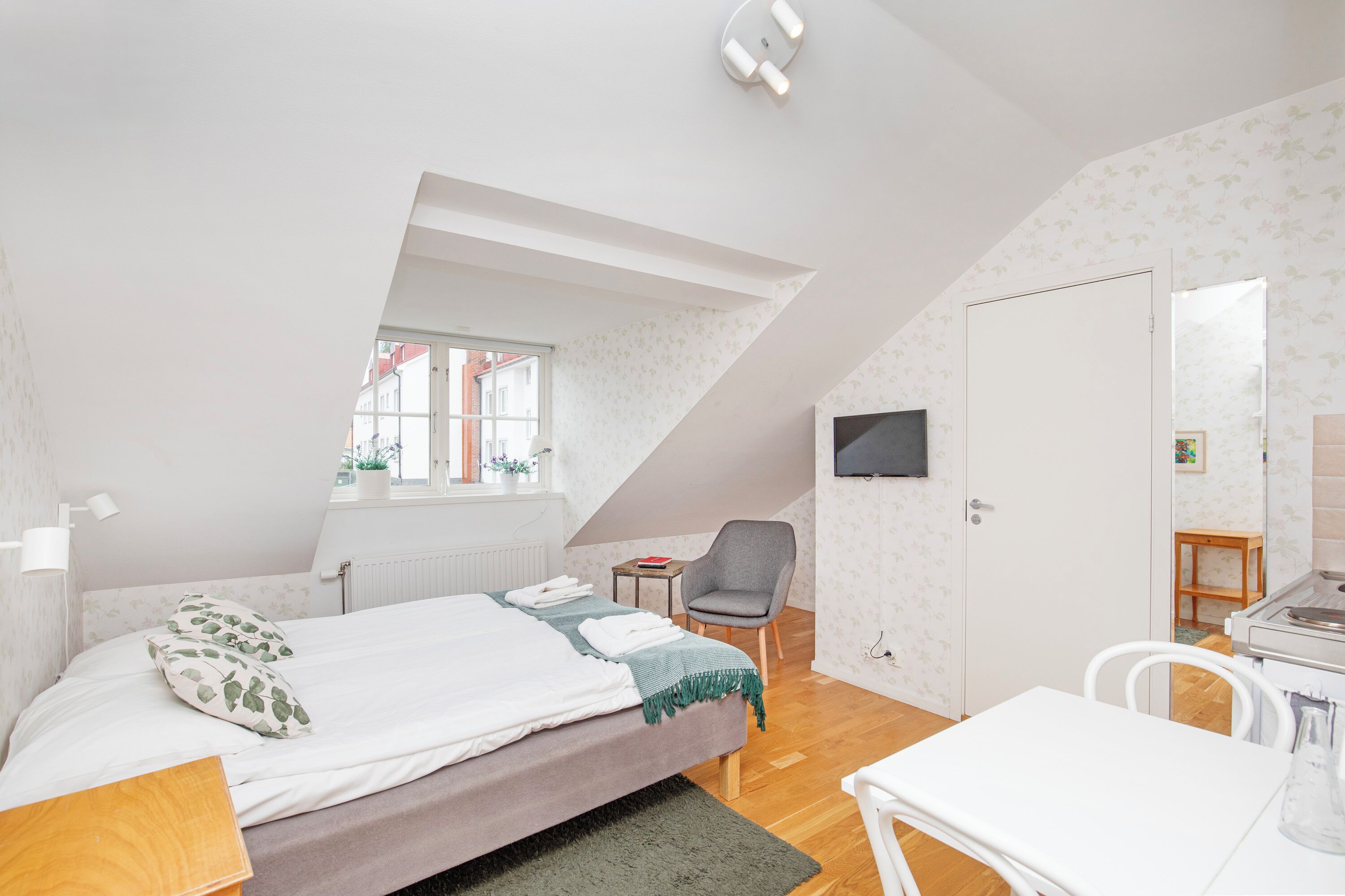 A bedroom with a bed, a desk, a chair, a TV, and a window.