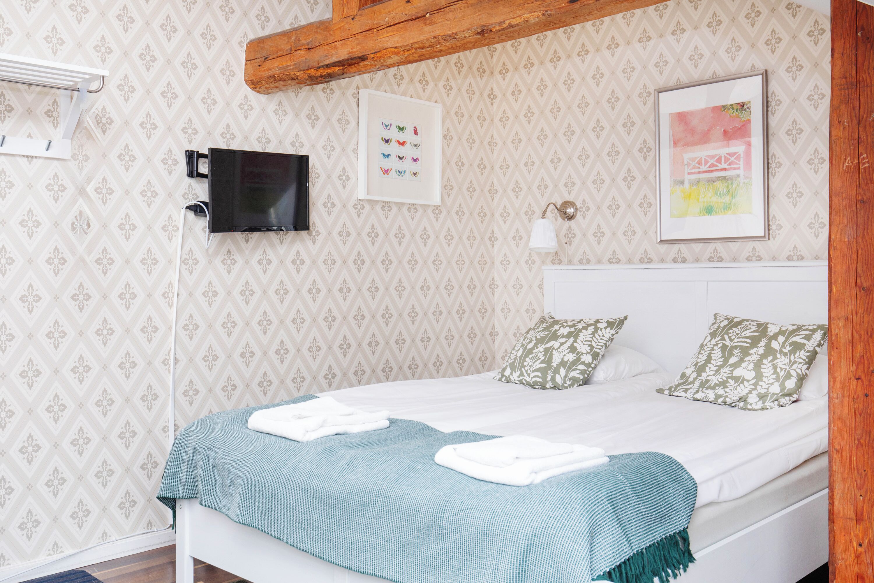 A neatly made bed with a blue blanket, a wall-mounted TV, and a framed picture on the wall.