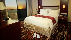 A hotel room with a large bed, a desk, a chair, a nightstand, and a view of the sunset. - Daamat Hotel (Addis Ababa)