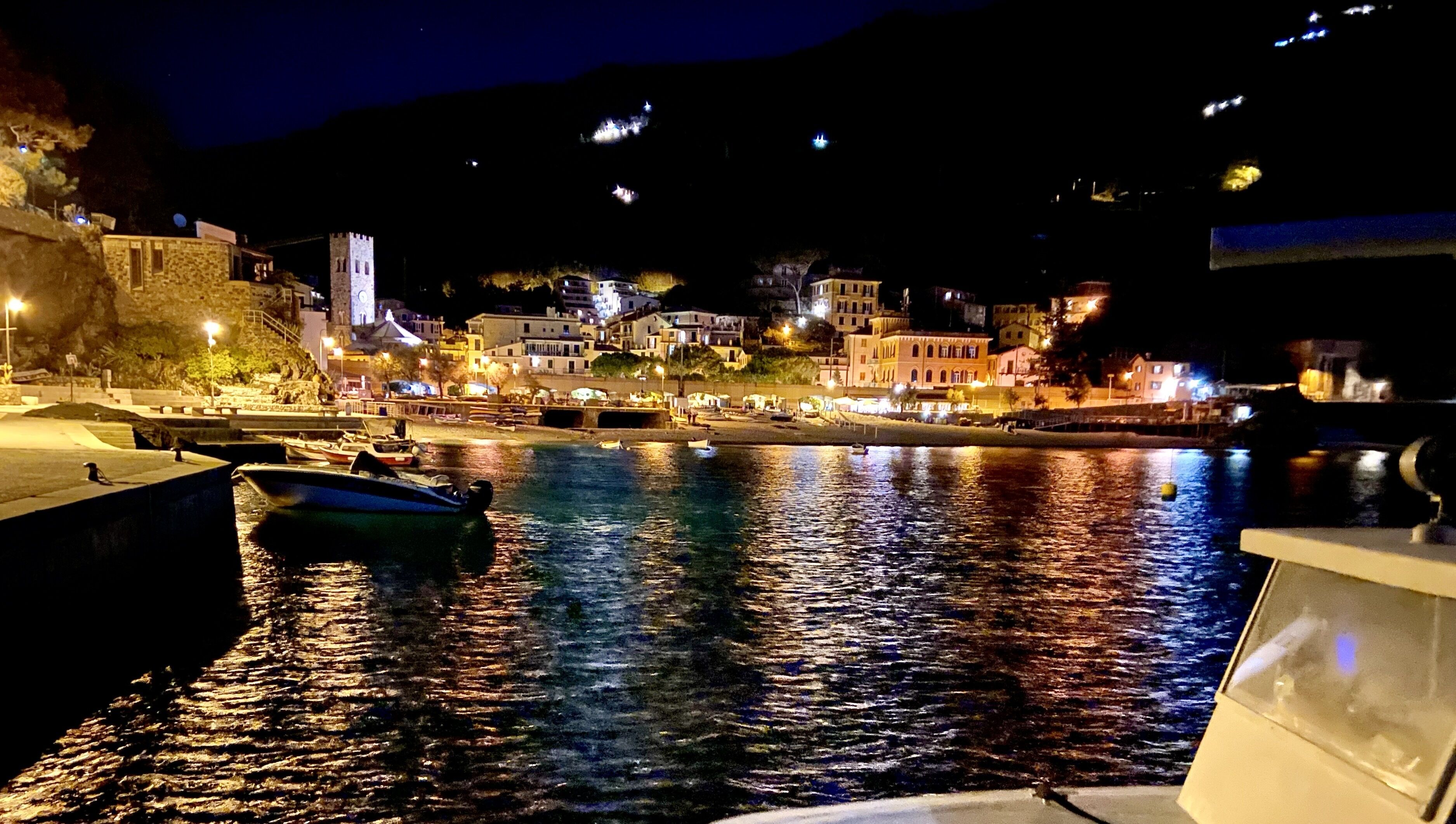 A coastal town at night with illuminated buildings, a stone tower, and boats docked at the harbor.