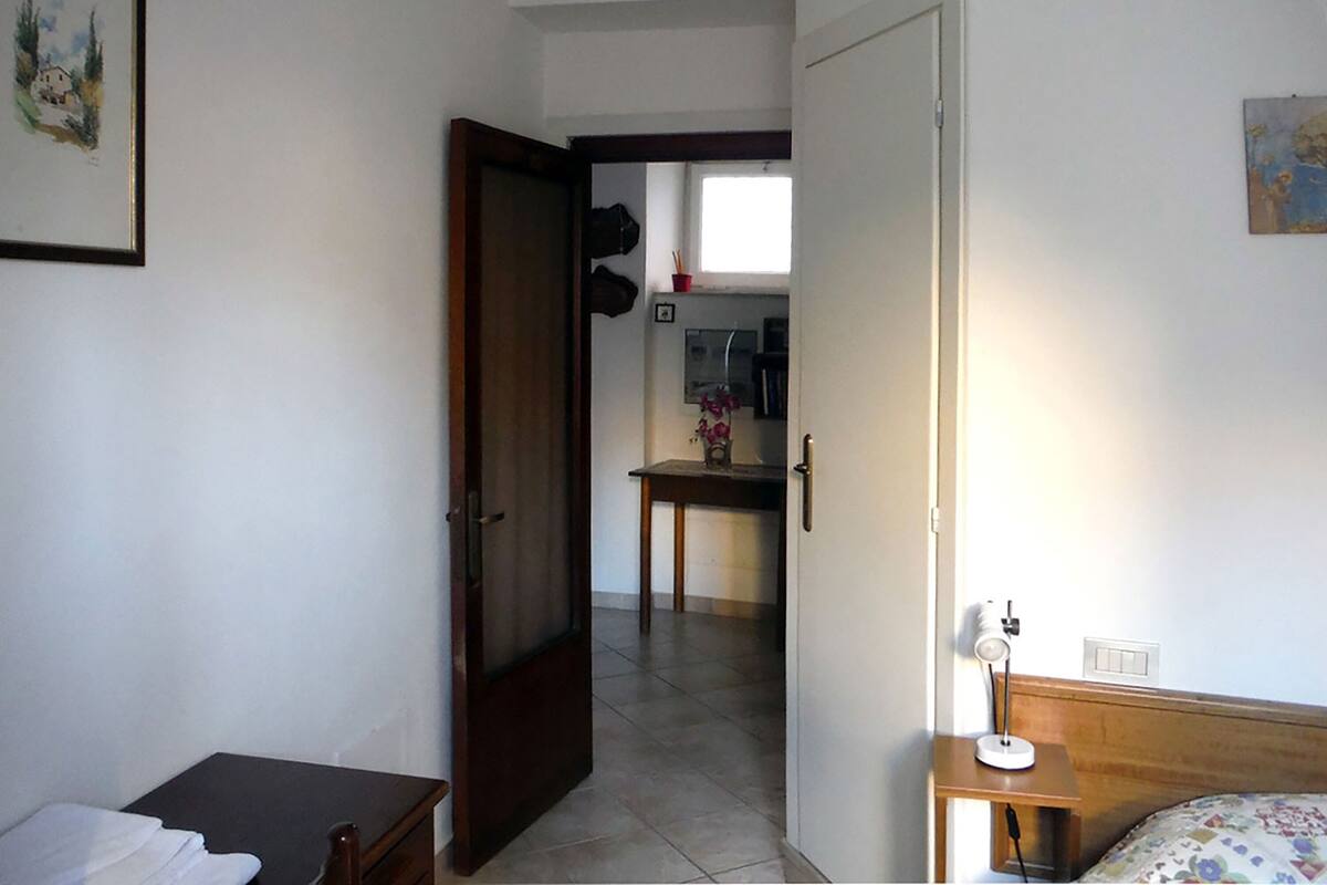 A bedroom with a bed, a bedside table, and a door leading to a kitchen area.