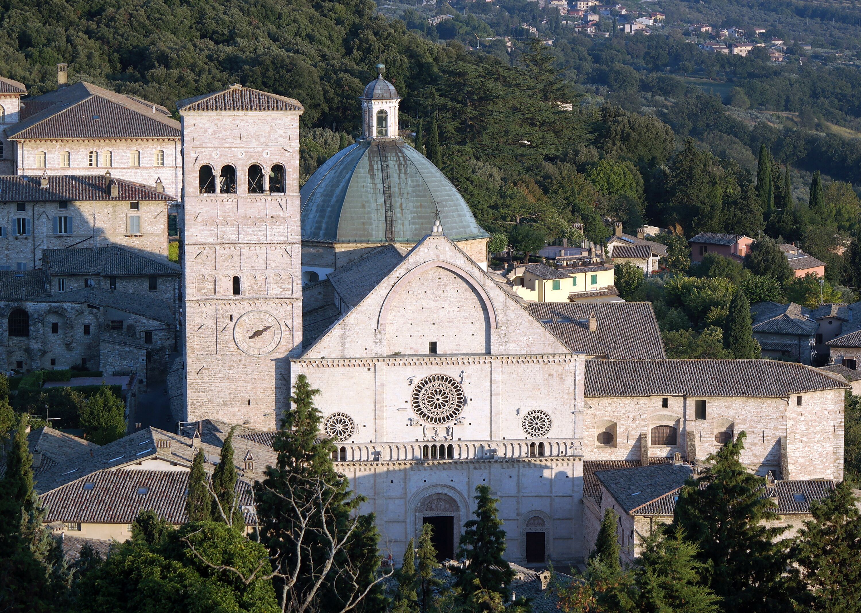 Uma igreja histórica com uma torre de sino imponente e uma grande cúpula, rodeada por edifícios tradicionais e vegetação.