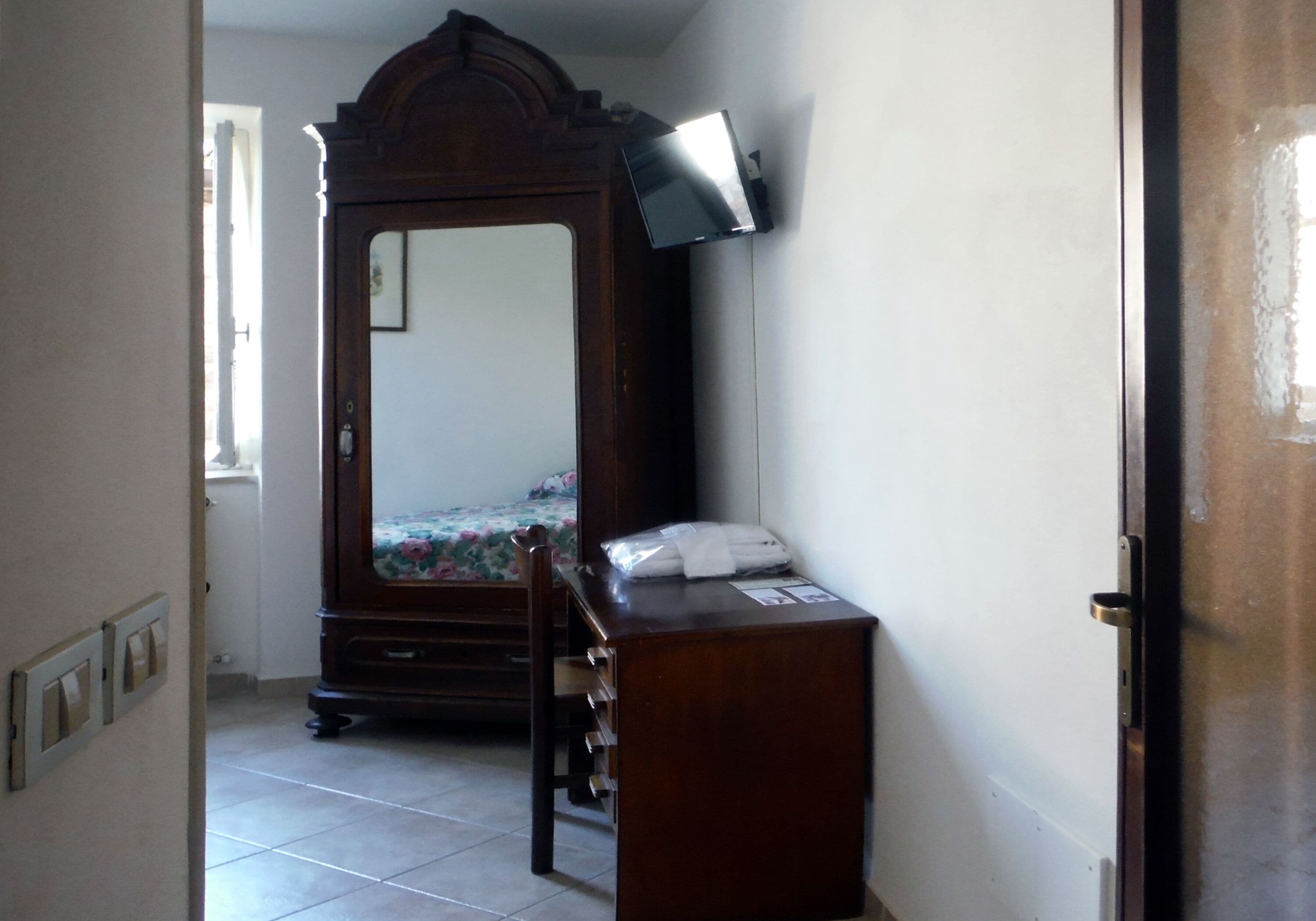 A bedroom with a wooden wardrobe, a bed with a floral bedspread, and a desk with a lamp.