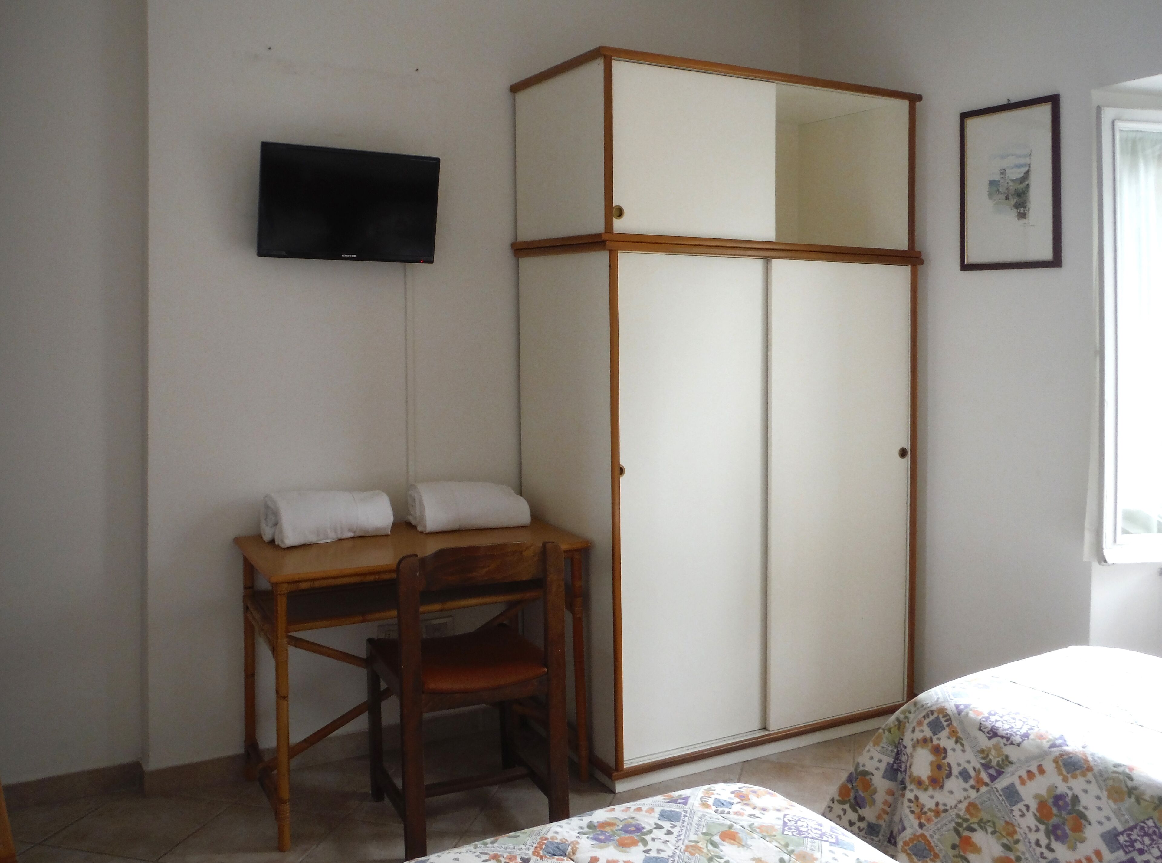 A room with a wooden desk, a chair, a wardrobe, a bed with a floral bedspread, a television mounted on the wall, and a framed picture.