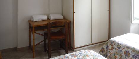 A room with a wooden desk, a chair, a wardrobe, a bed with a floral bedspread, a television mounted on the wall, and a framed picture.