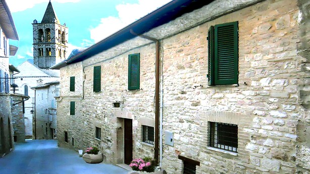 A narrow stone street with buildings featuring green shutters and a tower in the background. - Camere Gambacorta (Assisi)