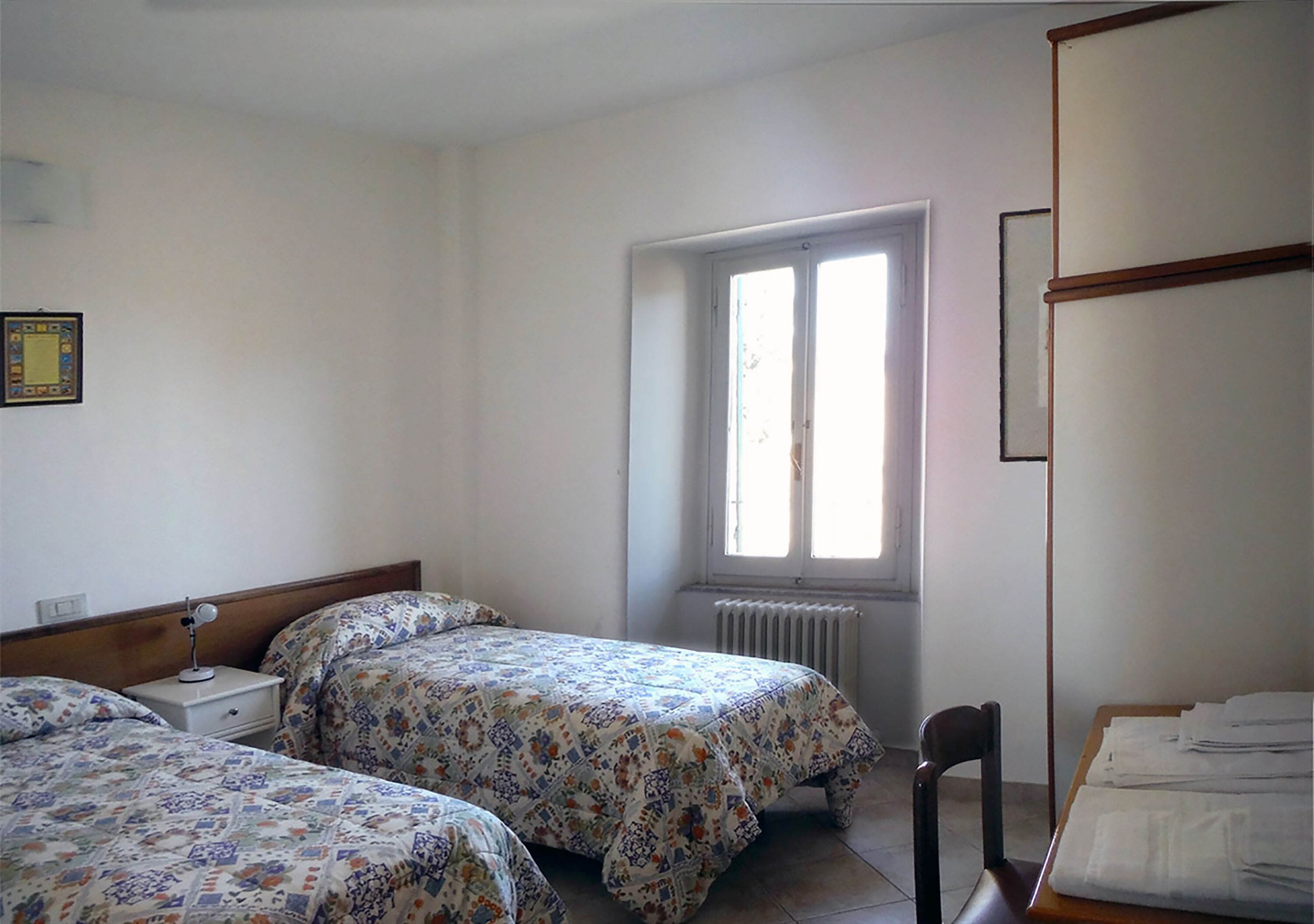 A bedroom with two beds, a radiator, a window, and a chair.