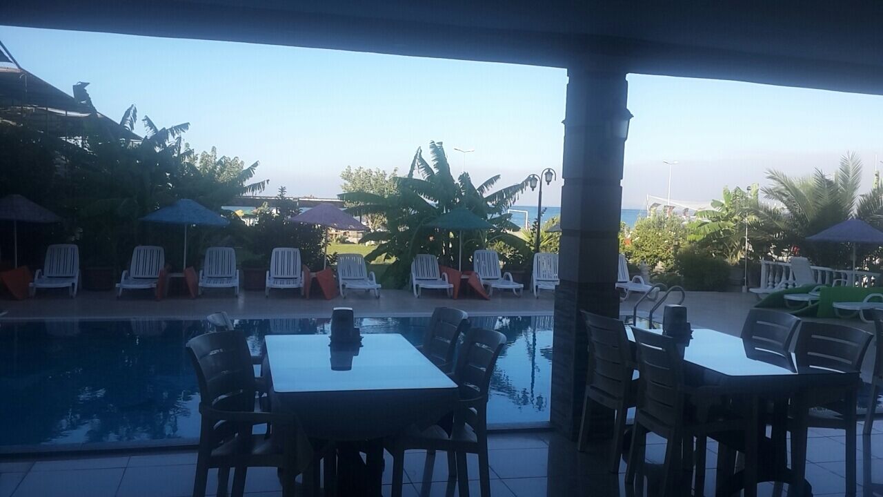 Un área junto a la piscina con sillas de descanso y mesas, rodeada de plantas tropicales y sombrillas.