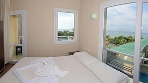 A hotel room with a bed, a window with a view of a pool and greenery, and a balcony with a railing. - Adahan Hotel (Menderes)