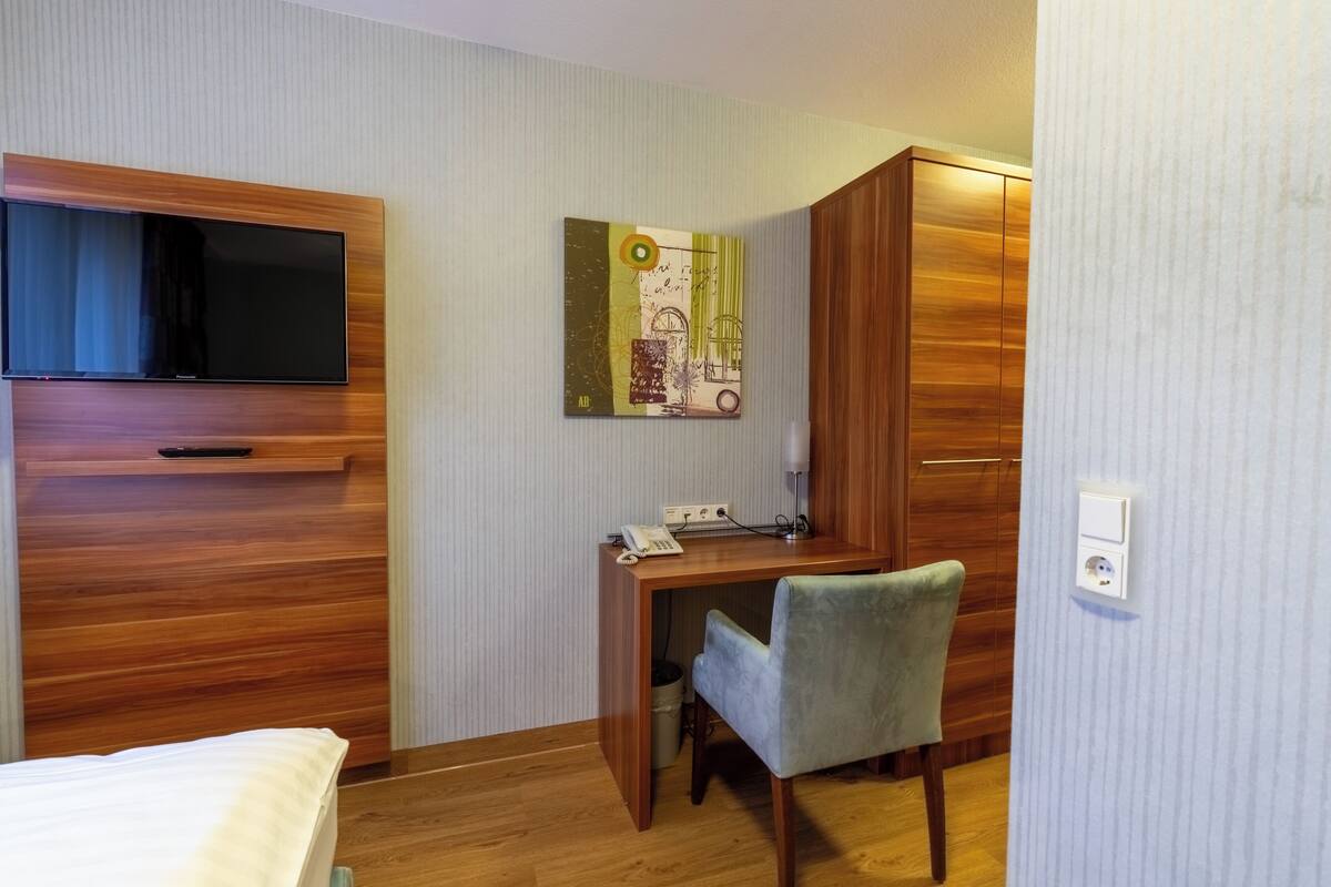 Habitación de hotel con cama, escritorio, silla, televisor montado en la pared y armario de madera.