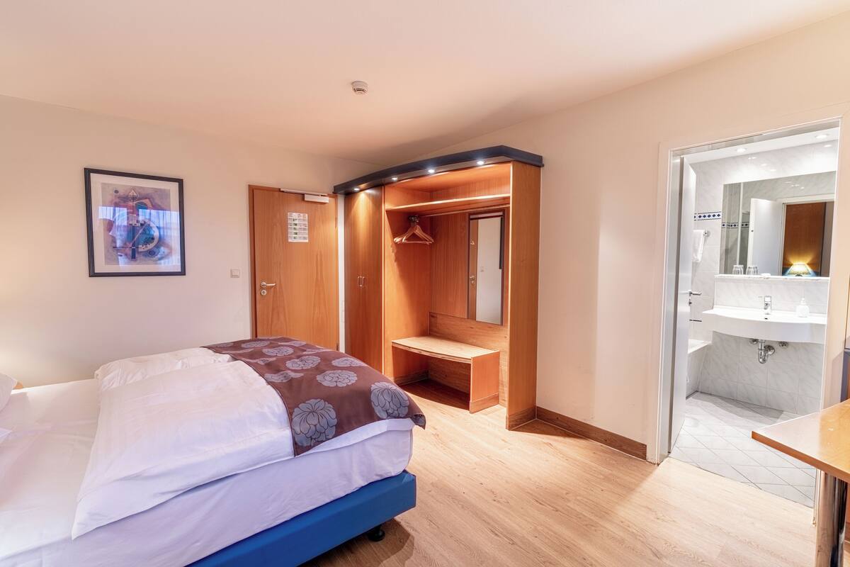 Un dormitorio con cama, armario, escritorio y baño con lavamanos y espejo.