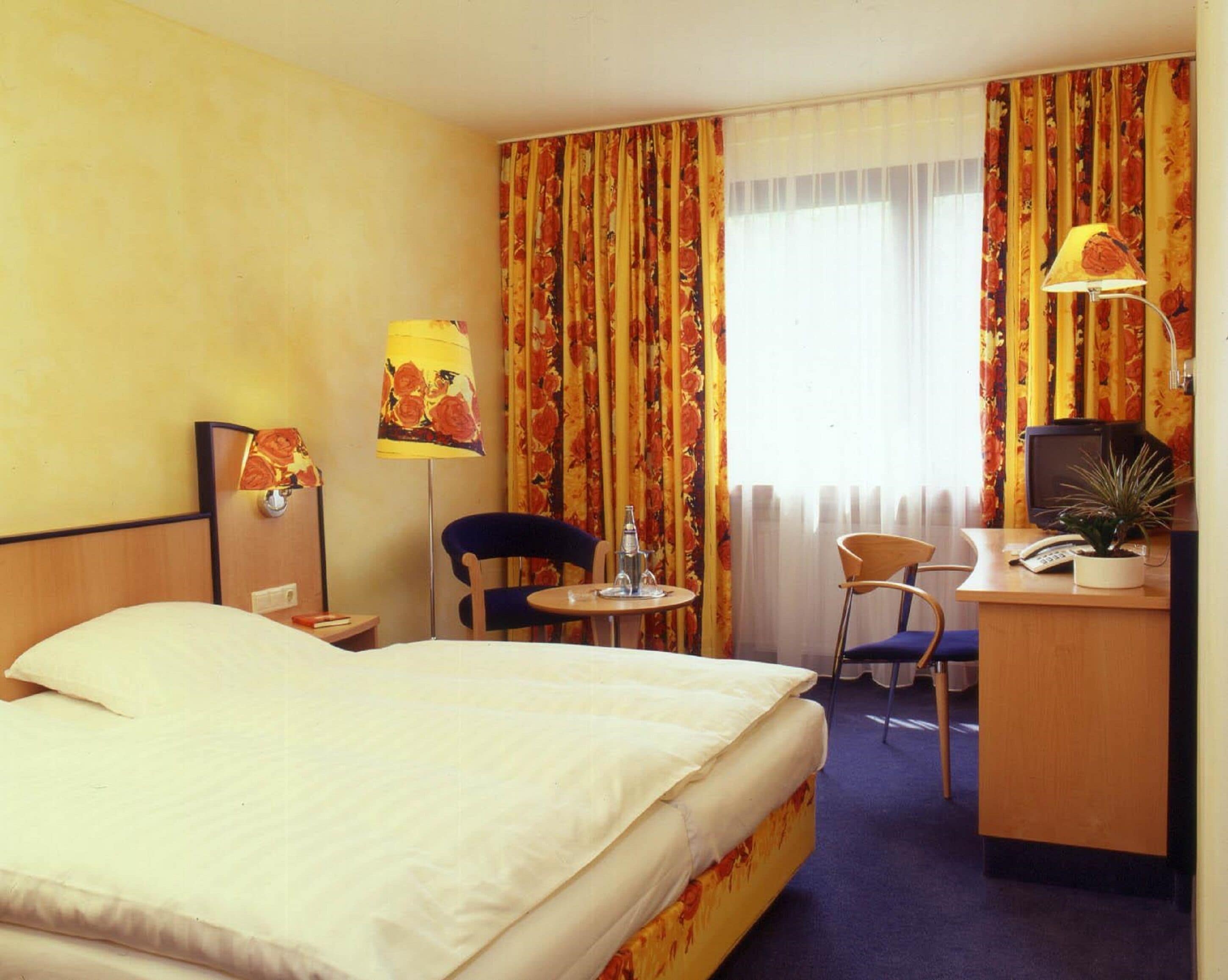 A hotel room with a bed, a desk with a computer, two chairs, a lamp, and a window with curtains.