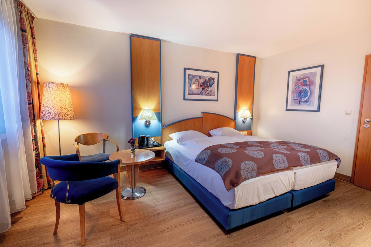 Una habitación de hotel con una cama, dos lámparas fijadas a la pared, una silla, una mesita y dos cuadros enmarcados.