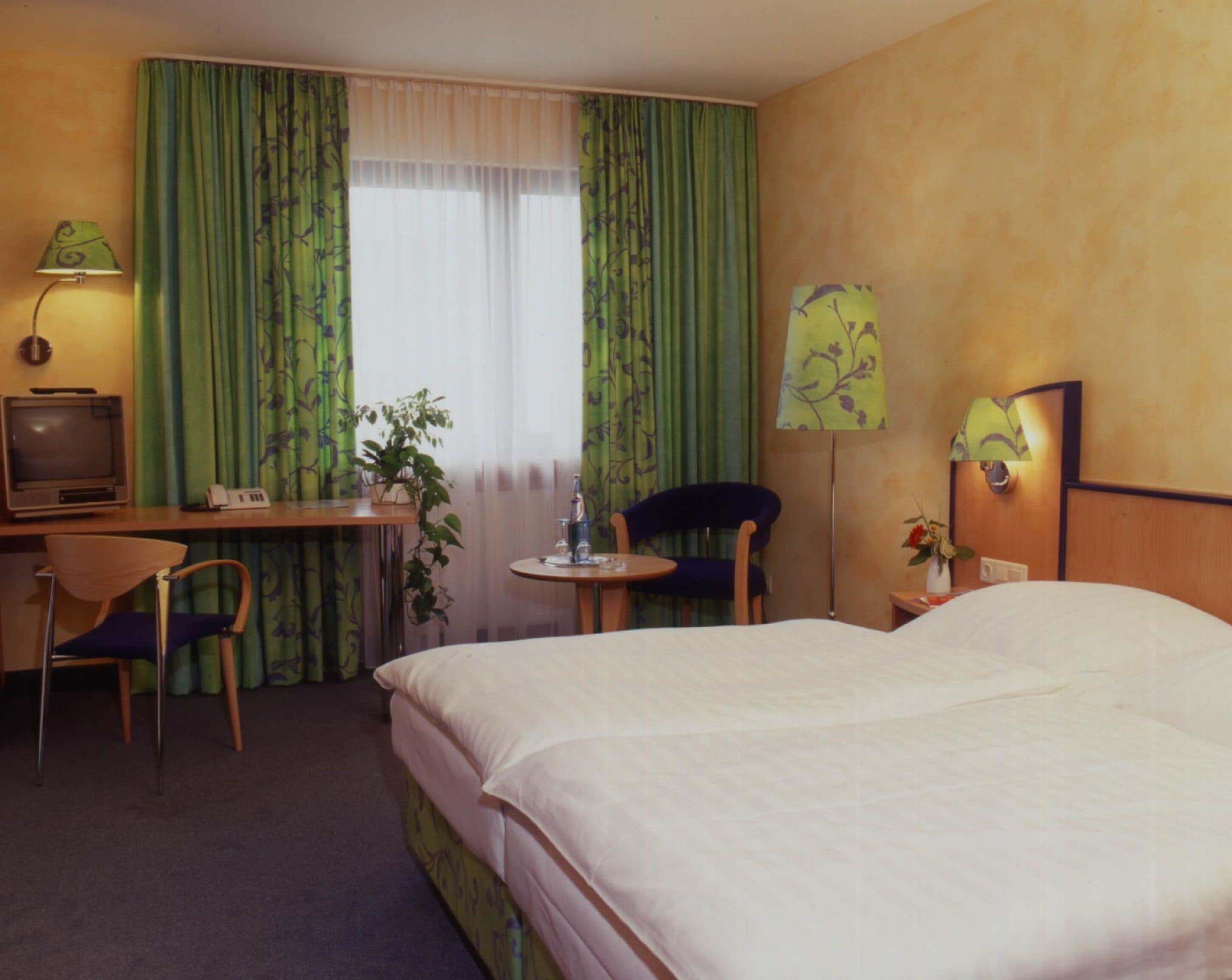 A hotel room with a bed, a desk with a computer and lamp, a chair, a small table with a vase, and a window with curtains.