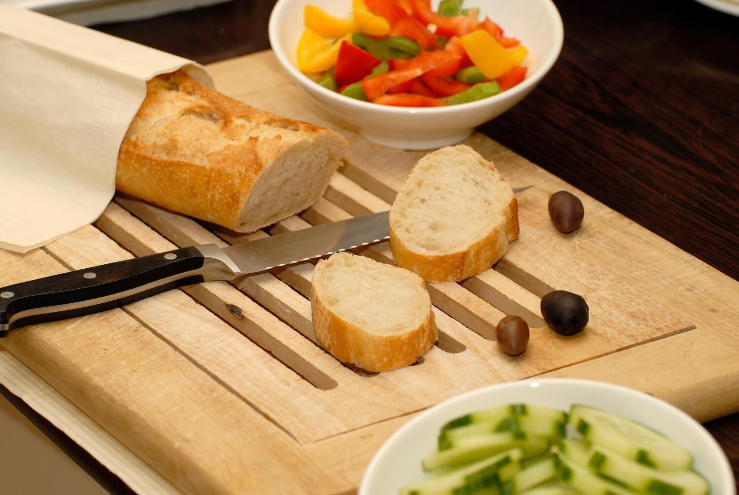 A loaf of bread on a wooden cutting board, sliced bread, a knife, and bowls of vegetables.