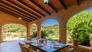 A covered outdoor dining area with a table set for four, surrounded by stone arches, overlooking a pool and lush greenery.