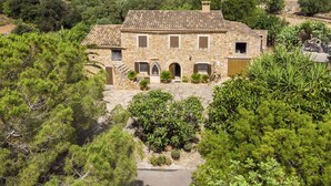 A stone house with a tiled roof, surrounded by dense greenery and hills in the background.