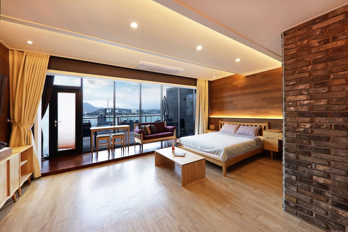 A modern hotel room with a large window, a bed, a seating area with a table, and a brick accent wall.
