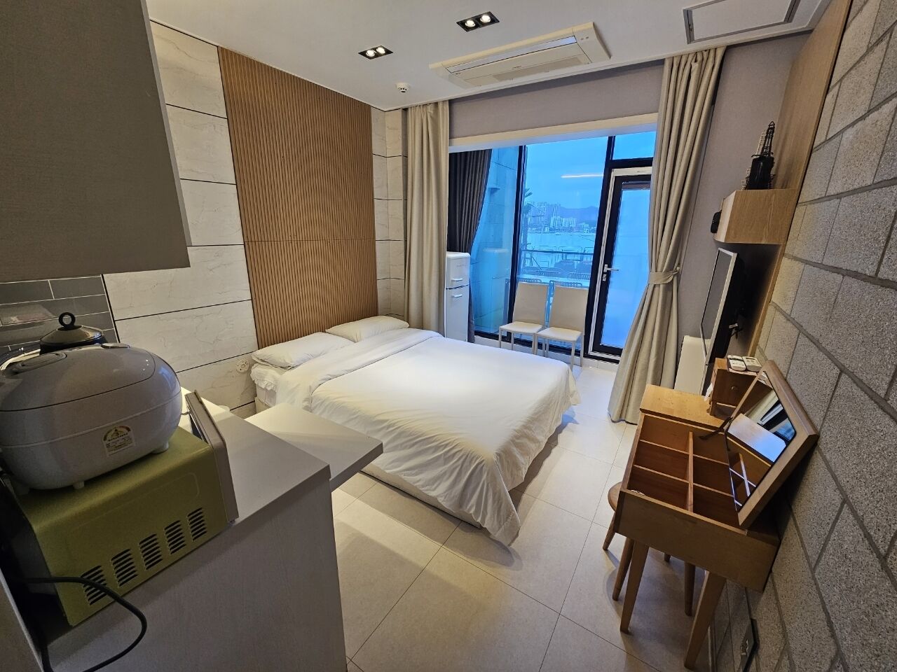 Quarto de hotel com cama, escrivaninha com computador, cadeira de massagem e vista da cidade por grandes janelas.