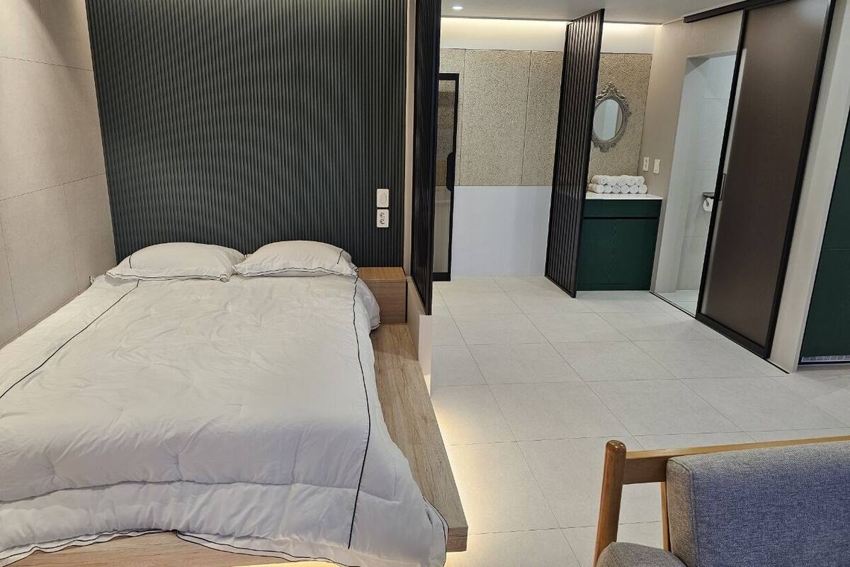 A modern bedroom with a large bed, a wooden nightstand, a chair, and a bathroom with a mirror and towel rack.