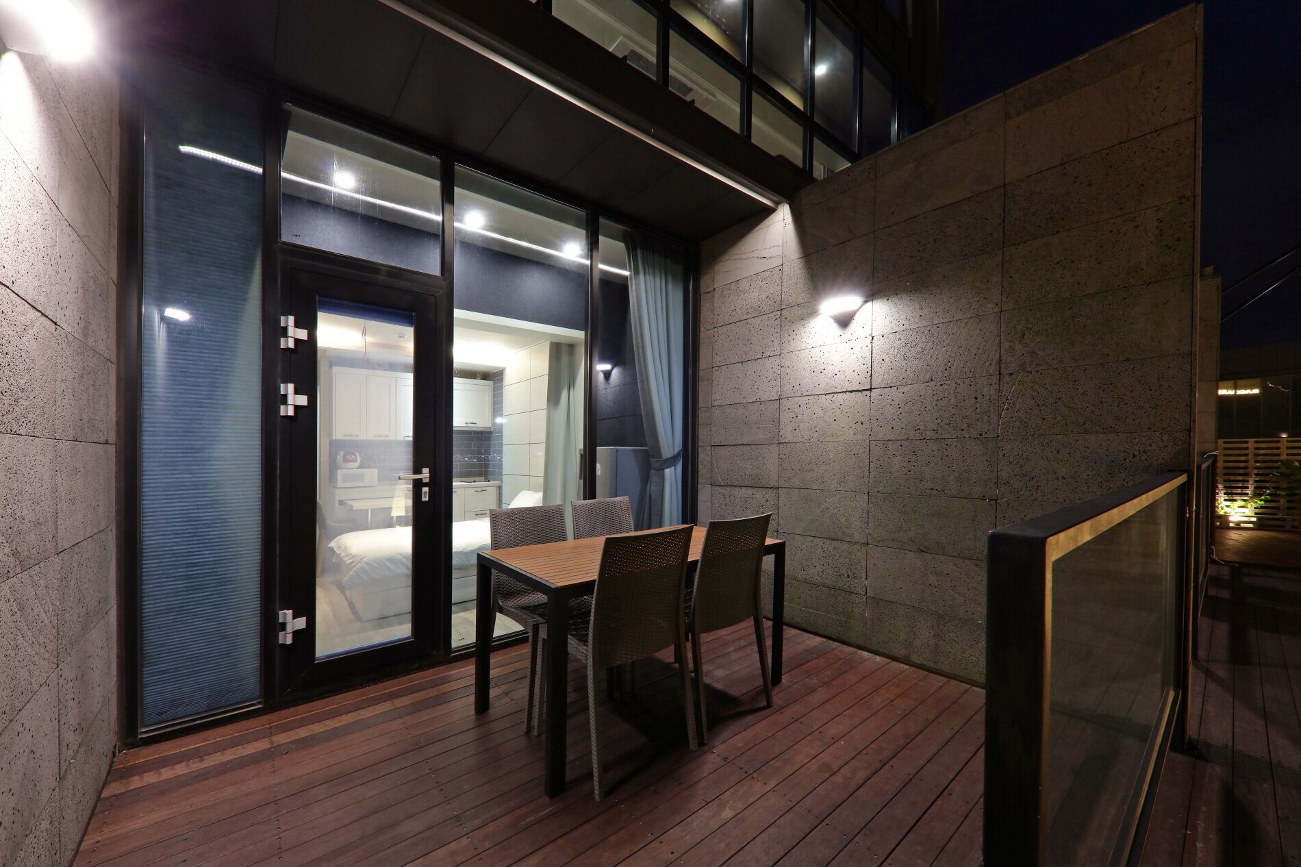 A modern hotel room with a wooden deck, a dining table with chairs, and a large window overlooking the city.