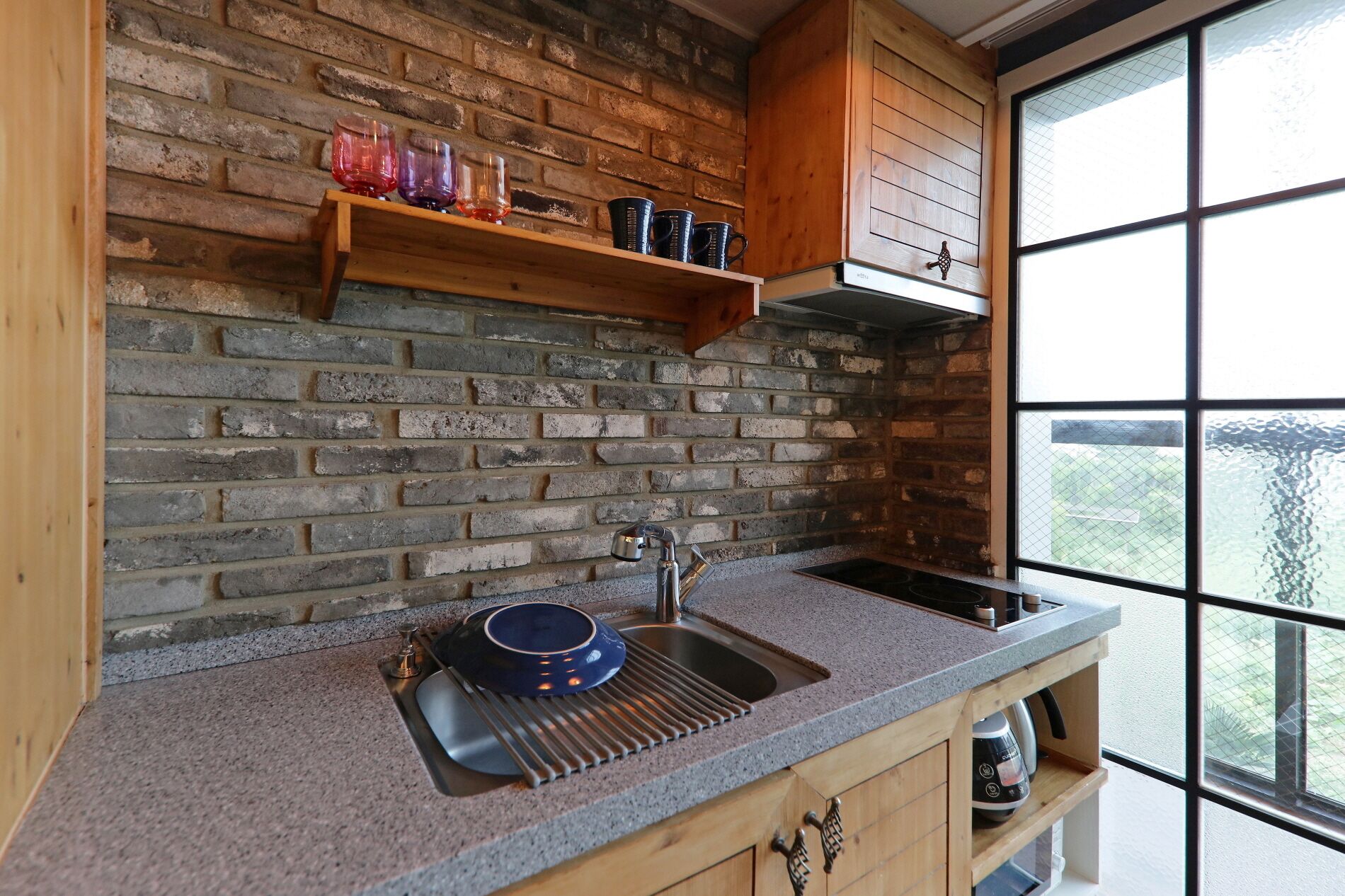A kitchen with a brick wall, wooden cabinets, a window, and a sink with a dish rack.
