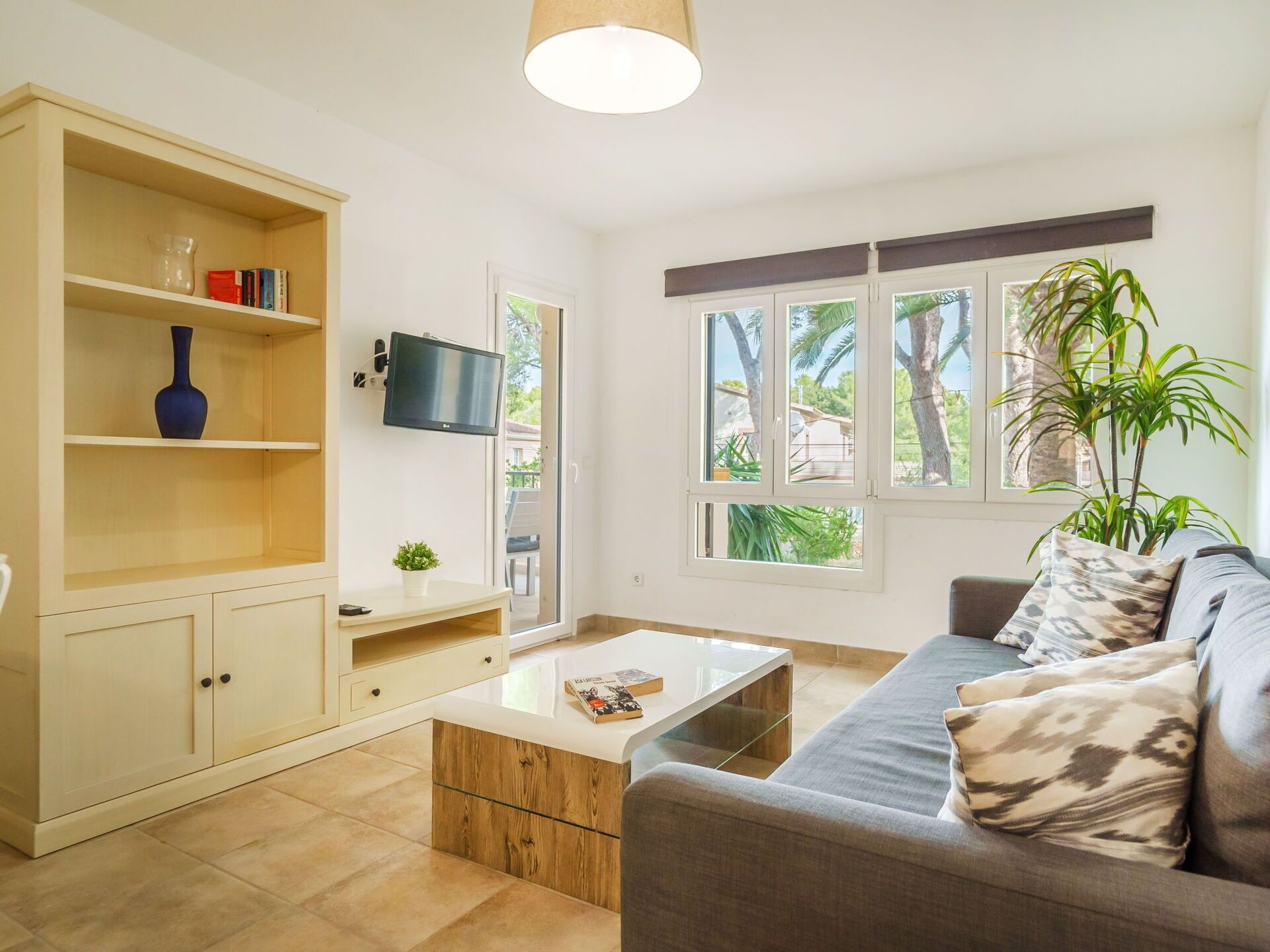 A living room with a sofa, coffee table, TV, bookshelf, and a view of palm trees outside.