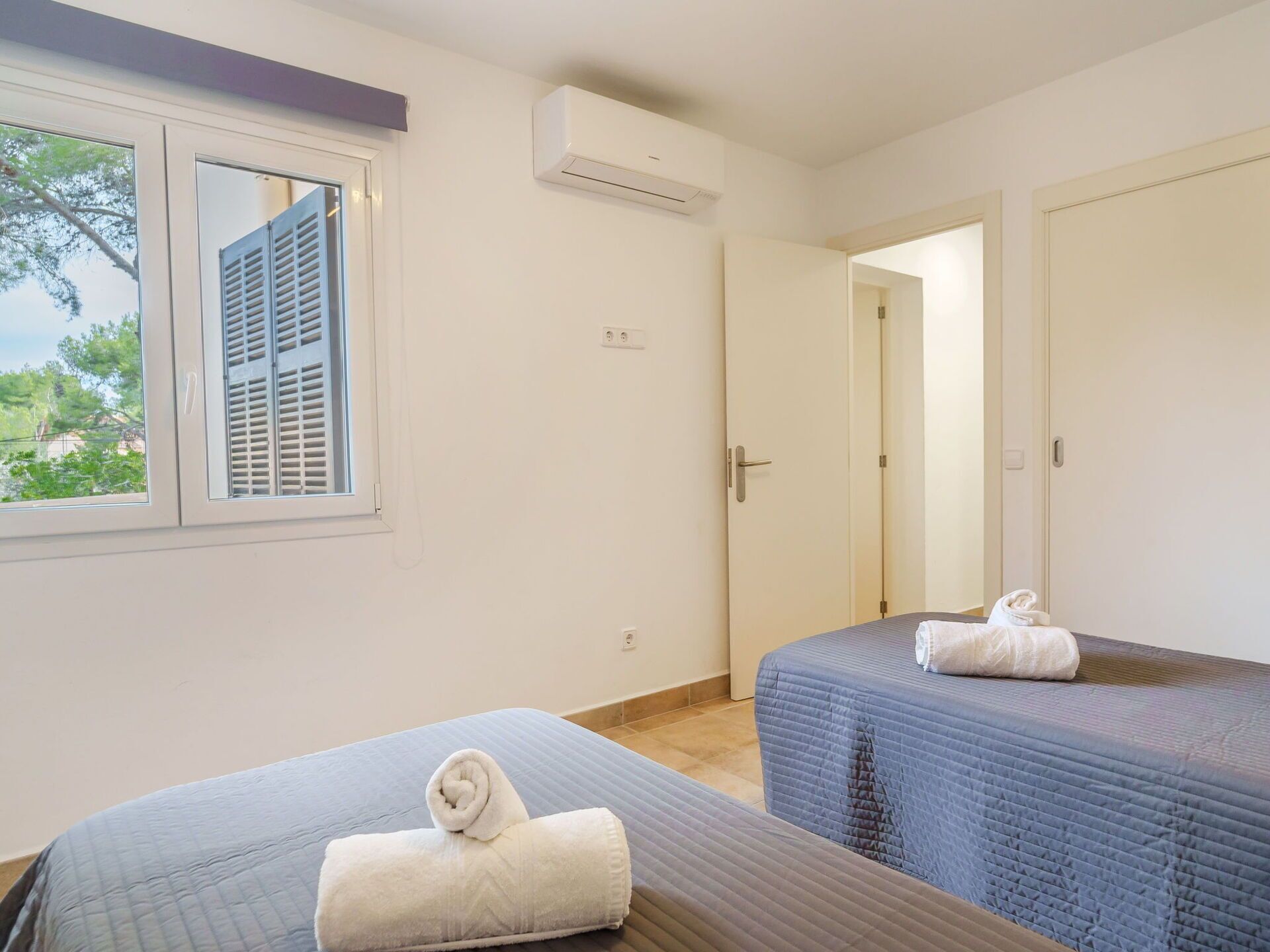 A hotel room with two beds, a window with shutters, an air conditioning unit, and a door leading to another room.