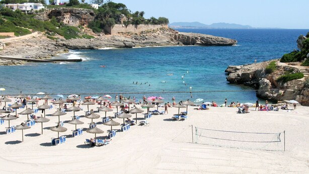 Une plage avec des transats et des parasols, un filet de volley-ball et, en arrière-plan, des falaises rocheuses.