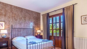 A bedroom with a metal bed frame, two bedside tables with lamps, a large window with curtains, and a radiator on the wall. - Nice apartment for 4 guests with pool, WIFI, TV and terrace (Manacor)