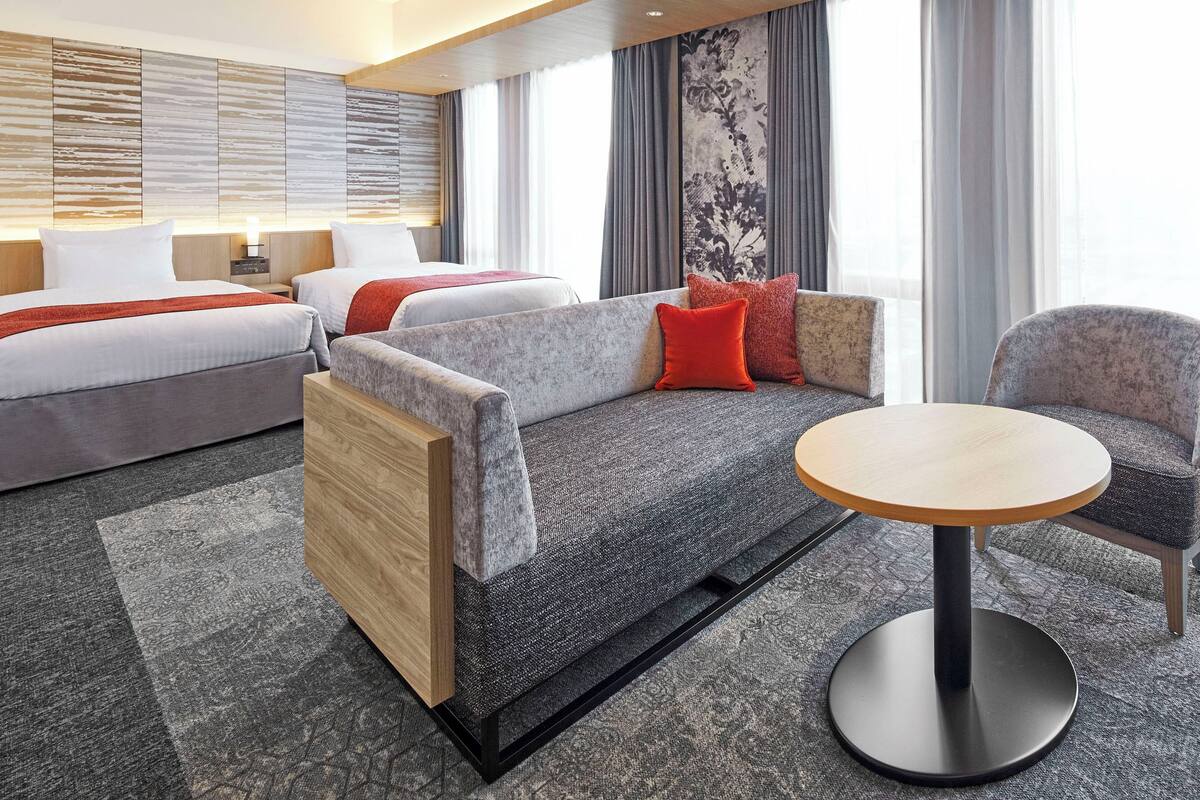 Une chambre d’hôtel moderne avec un canapé, deux lits, une table ronde et une grande fenêtre avec des rideaux.
