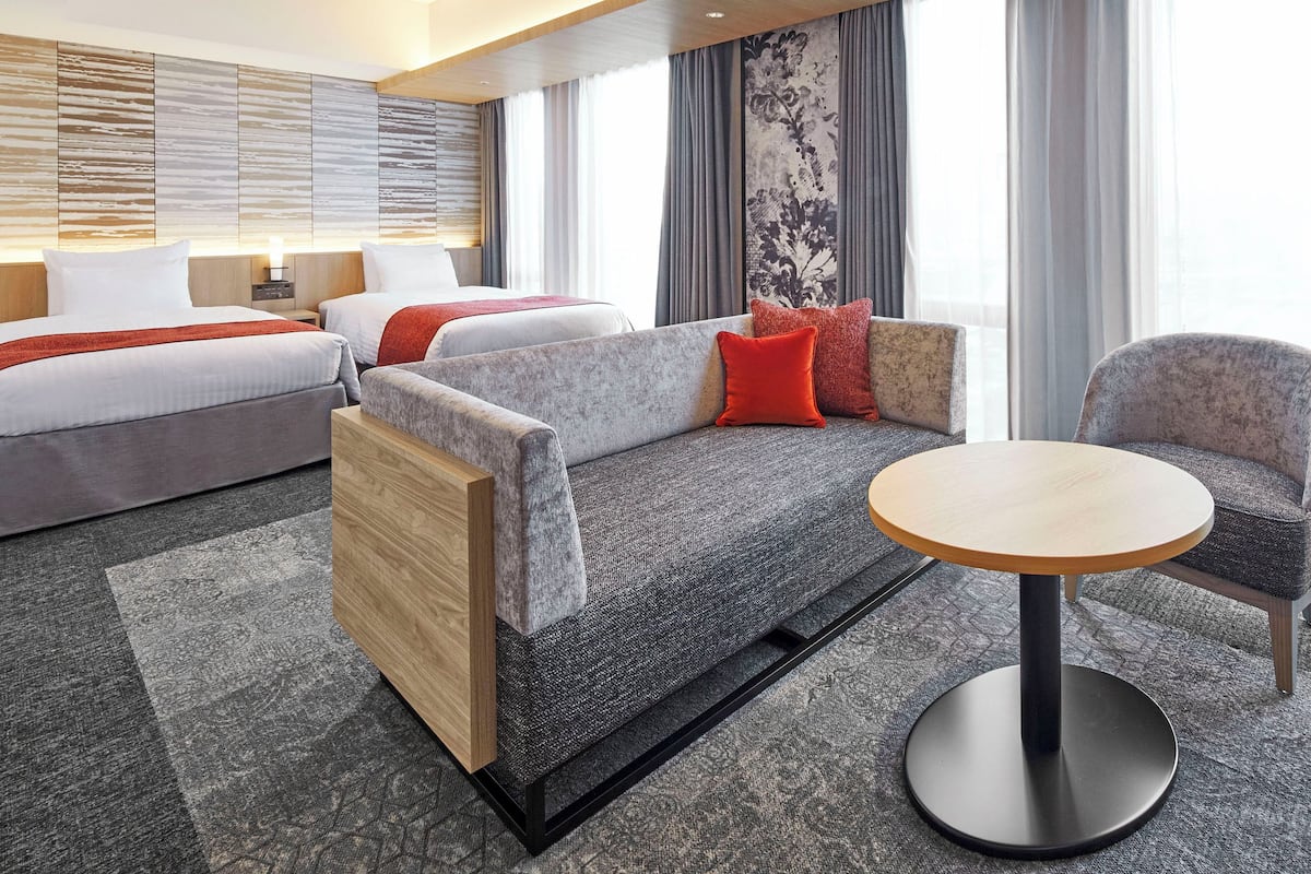 Une chambre d’hôtel moderne avec un canapé, deux lits, une table ronde et une grande fenêtre avec des rideaux.