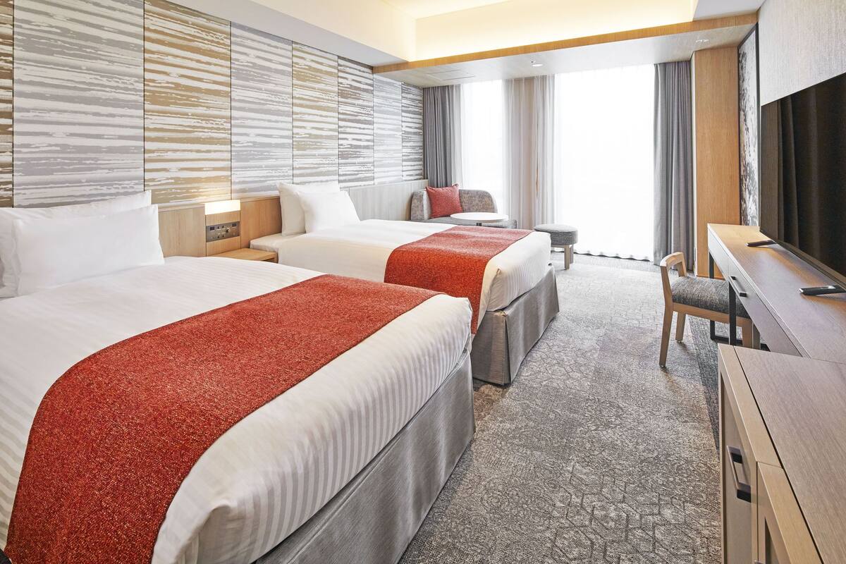 Une chambre d’hôtel avec deux lits, un bureau, une chaise, une télévision et une grande fenêtre avec des rideaux.