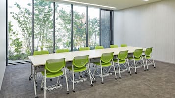 A conference room with a long table and green chairs, large windows with a view of trees, and a carpeted floor.