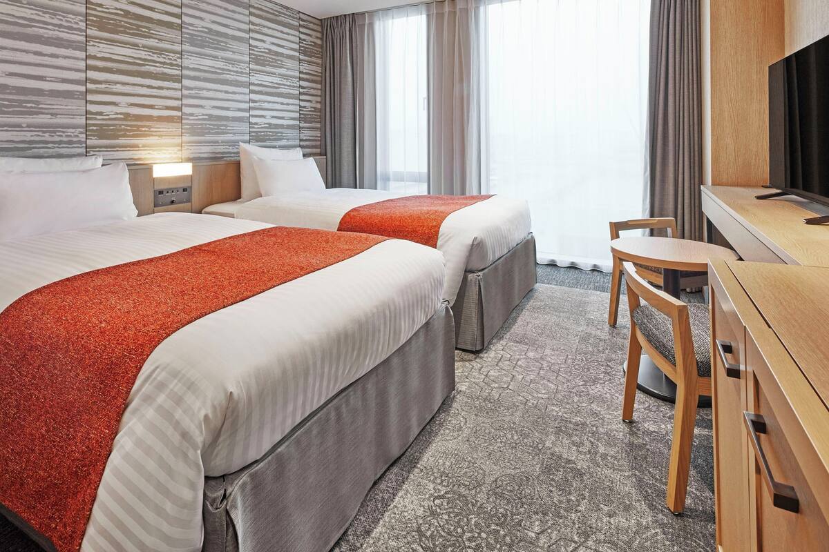 Une chambre d’hôtel avec deux lits, un bureau, une chaise, une télévision et une grande fenêtre avec des rideaux.
