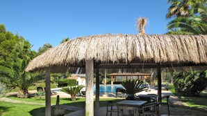 Un pabellón con techo de paja, asientos al aire libre, piscina y abundante vegetación.