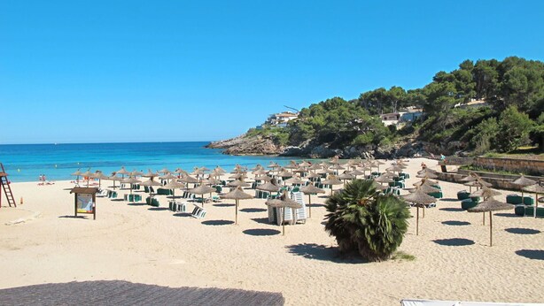 Une plage dotée de nombreuses chaises longues et de parasols, d’une terrasse en bois, et offrant une vue sur la mer et une colline parsemée de bâtiments.