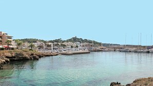 Un port de plaisance avec des bateaux amarrés, une eau claire, une côte rocheuse et des bâtiments en arrière-plan.