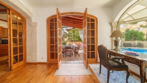 Une pièce spacieuse dotée d’un plancher en bois, d’un bureau avec une chaise et d’une vue sur une piscine, visible à travers une fenêtre en arc de cercle.