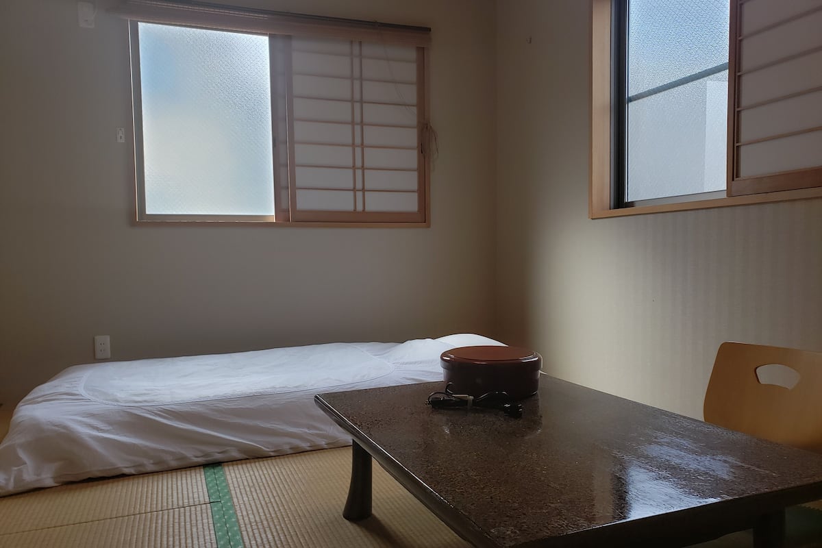 A small room with a bed, a table, and a chair. There is a window with a view of the sky.