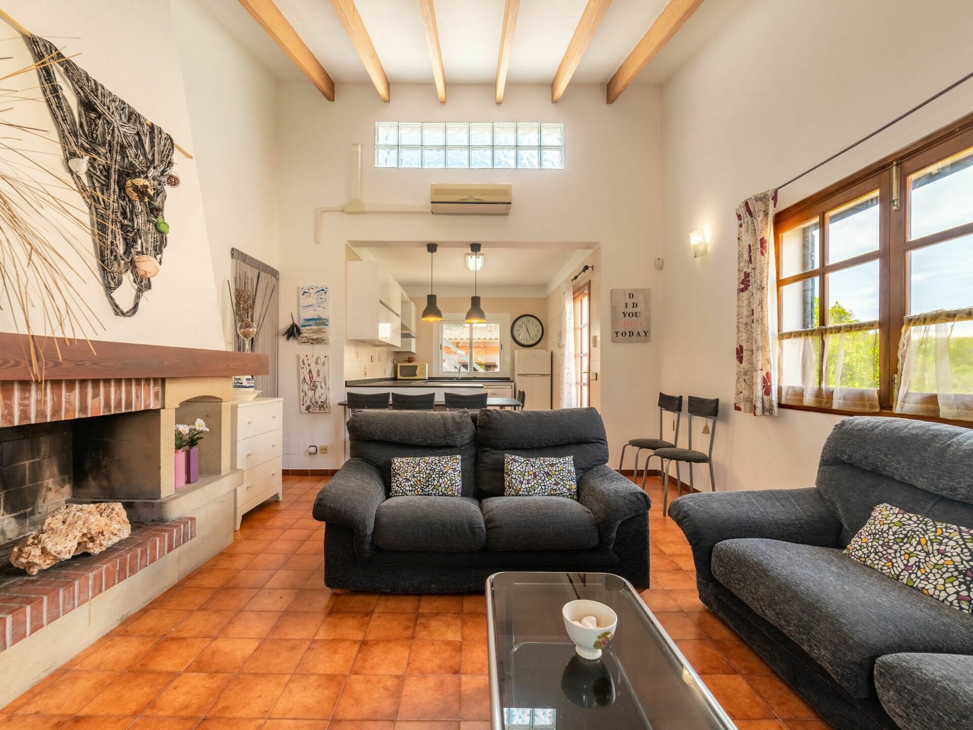 A spacious living room with a fireplace, two grey sofas with patterned cushions, a coffee table, and a view of the kitchen and dining area through a large window.